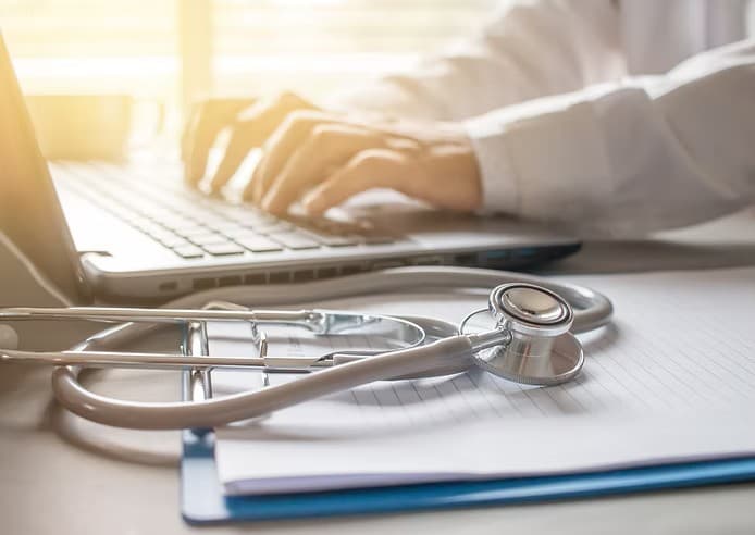 Close-up of a stethoscope on a clipboard with a doctor typing on a laptop.